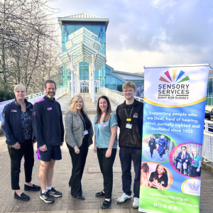 Kathy and Ben stand at the gangway to Guildford Spectrum's white and glass leisure centre building with three staff members from Guildford Spectrum, next to a Sensory Services by Sight for Surrey pop-up banner supporting people who are Deaf, hard of hearing, blind, partially sighted and deafblind. White text within pink bubble at top Guildford Spectrum’s Charity for 2026!