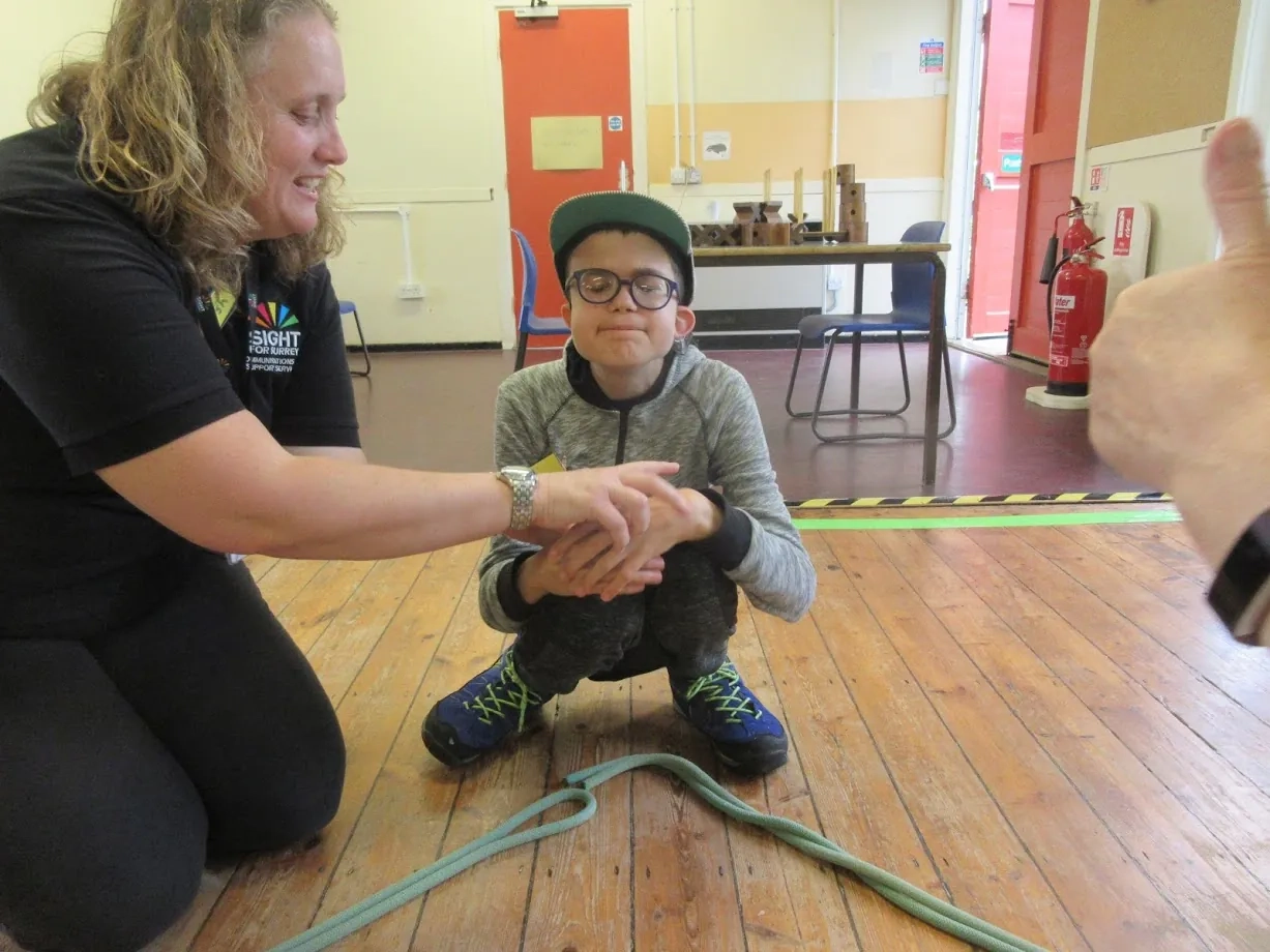 A young vision impaired boy is squatting on the floor and a member of staff is holding his hands