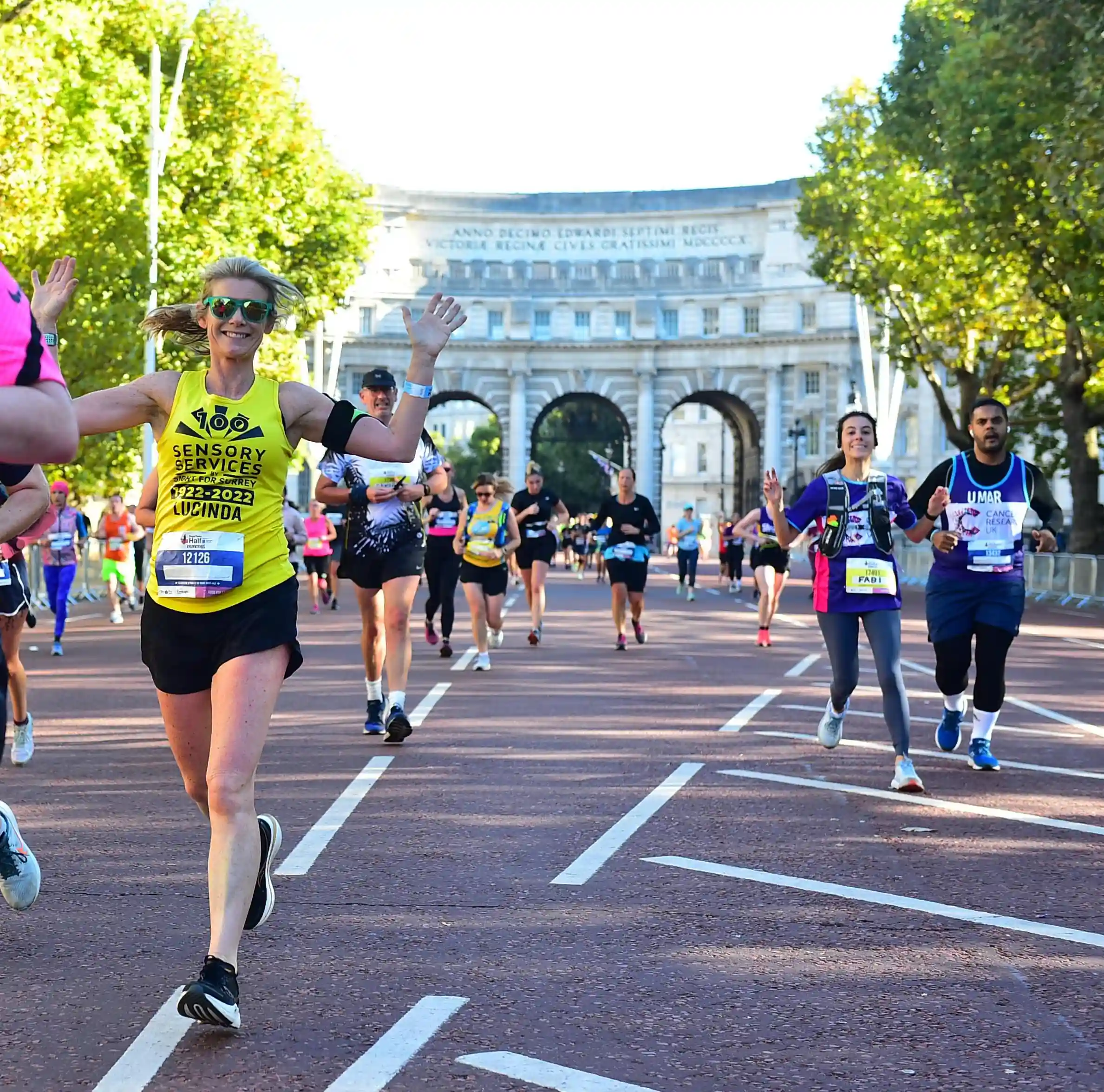 A runner wearing a branded Sensory Services by Sight for Surrey t-shirt with Pal Mall behind her, she is running towards the camera waving her arms, other runners are around her.