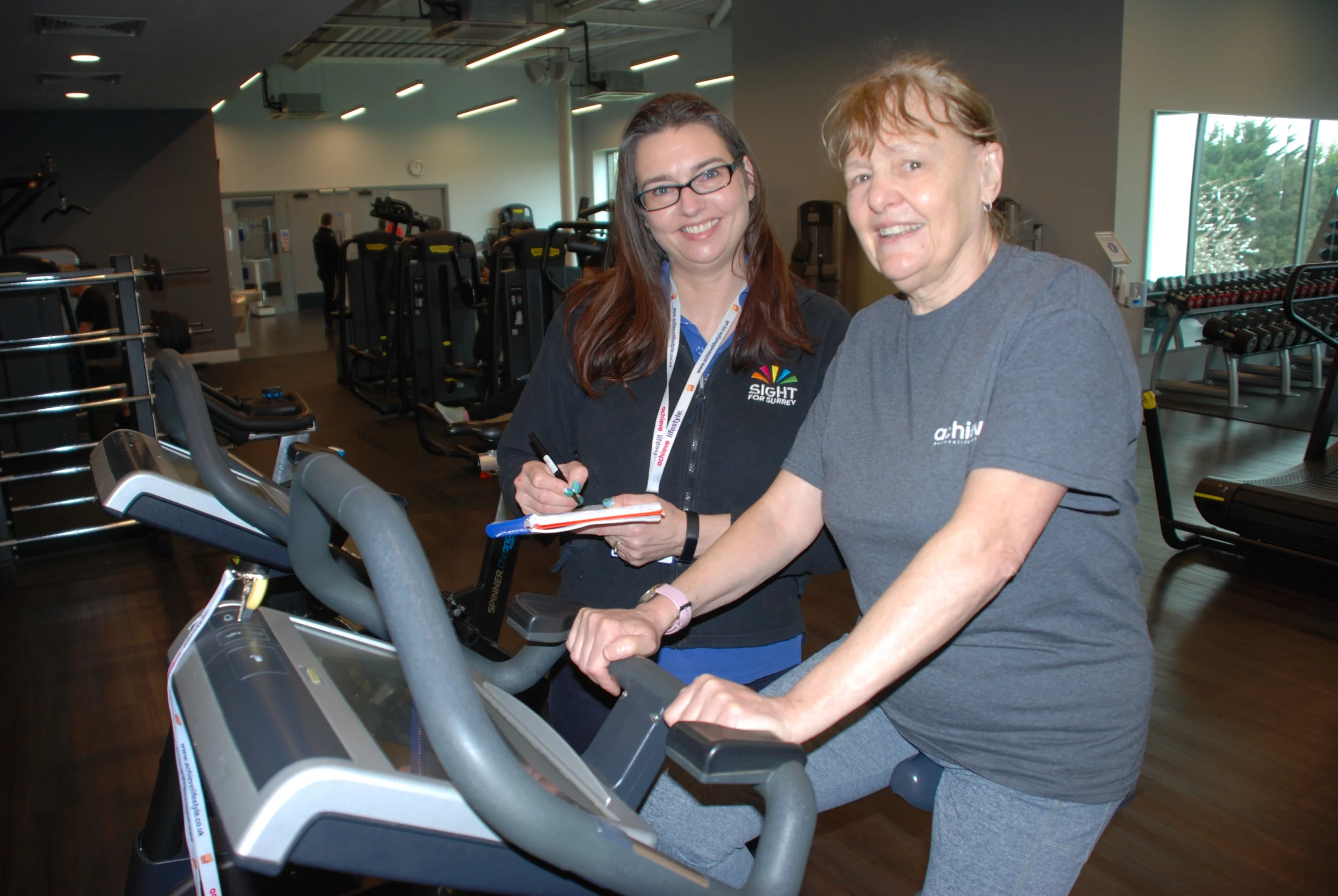 Larraine with Rehabilitation Support Worker, Suzie, at the gym. Larraine is on an exercise bike and Suzie is standing by her side.