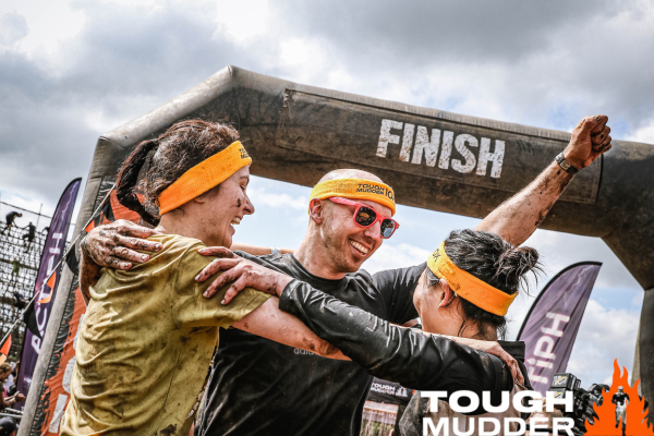 The finish line at a Tough Mudder event, with three very muddy people hugging each other.