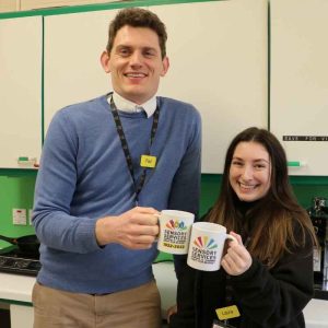 Two members of staff are smiling at the camera holding branded charity mugs in their hands