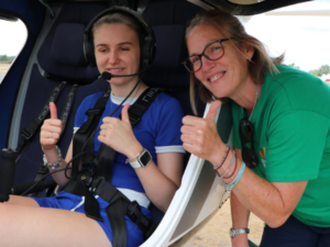 A young girl we support sitting in the plane with in-flight headphones on and standing beside her is Carole from the children and young people's service
