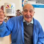 A man is holding a mug aloft in a cheers pose, he is elderly and smiling at the camera.