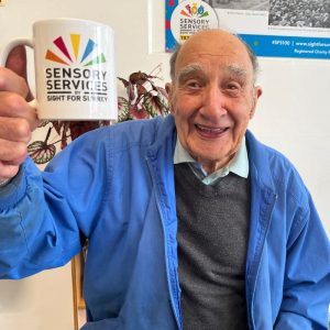 A man is holding a mug aloft in a cheers pose, he is elderly and smiling at the camera.