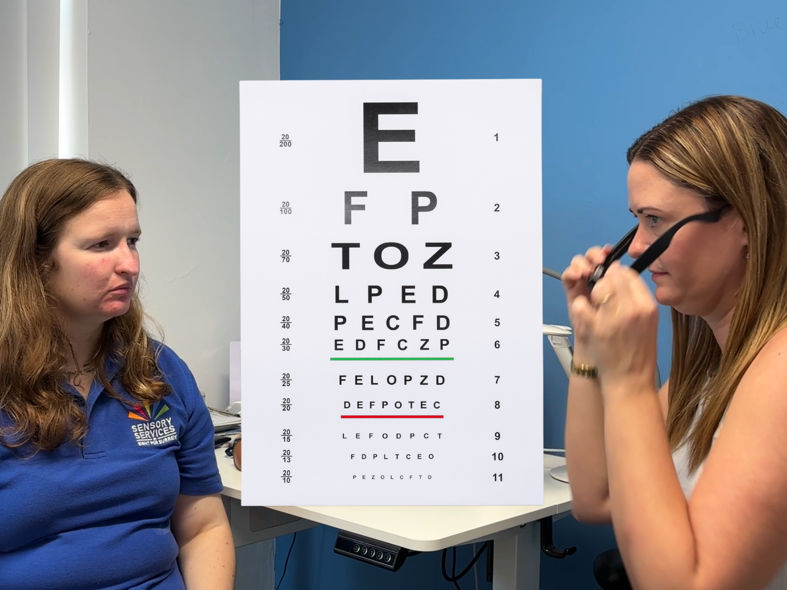 A woman is trying on glasses at an eye test as the optician in blue polo shirt looks on. An eye chart is central.